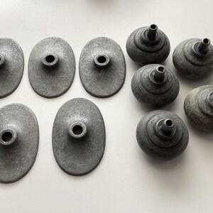 May include: A collection of eight antique metal hardware pieces. The set includes five oval-shaped backplates and three round, knob-like components, all in a weathered grey finish. These pieces appear to be for furniture or cabinetry.