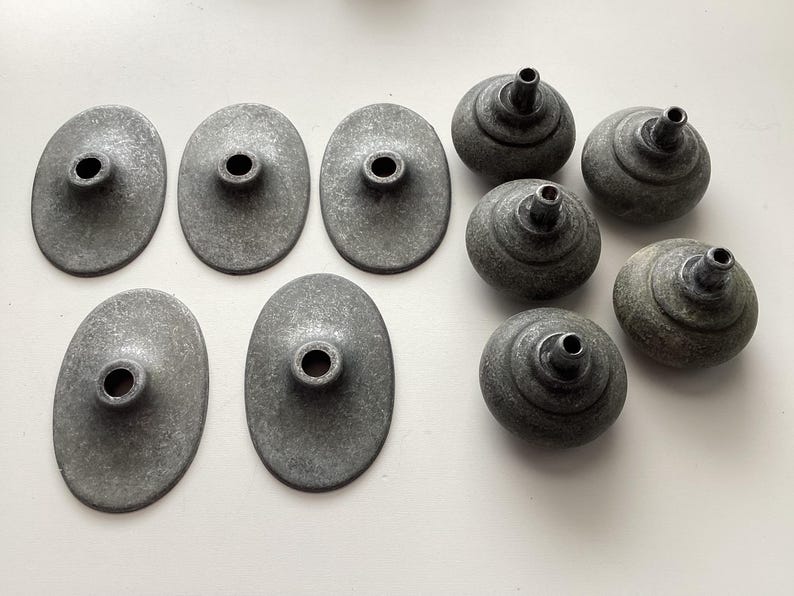 May include: A collection of eight antique metal hardware pieces. The set includes five oval-shaped backplates and three round, knob-like components, all in a weathered grey finish. These pieces appear to be for furniture or cabinetry.