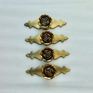 May include: Four antique brass drawer pulls. Each pull features a rose-shaped knob centered on a decorative, curved backplate. The brass has a warm, aged finish, suggesting vintage or classic design. Ideal for furniture restoration or adding a touch of elegance.