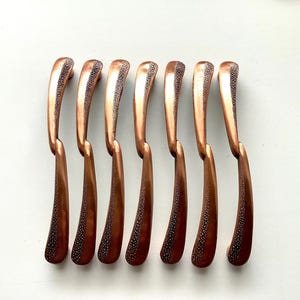 May include: Seven copper-colored cabinet pulls with a curved design. The pulls have a textured, dotted pattern on the handle and a smooth, curved shape. The hardware is arranged in a row against a white background.