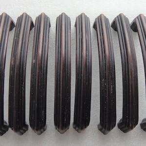 May include: Seven dark bronze cabinet pulls with a vertical ribbed design. The pulls have a curved shape and are arranged in a row. The finish has a slightly distressed appearance, adding a vintage aesthetic. These pulls are suitable for drawers or cabinet doors.