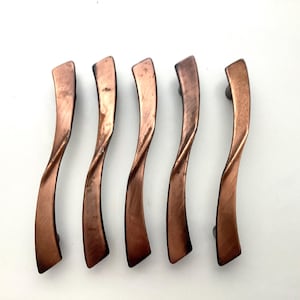 May include: Six copper-colored cabinet pulls with a twisted design. Each pull has a curved shape and a dark finish, with a mounting post on each end. The pulls are arranged on a white background.