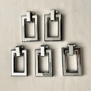 May include: Five shiny, rectangular cabinet pulls with a modern design. Each pull features a rectangular frame with a raised, rectangular top piece. The pulls are made of a reflective, silver-colored metal.