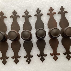 May include: Six antique bronze cabinet knobs and pulls. Each pull has a decorative, elongated backplate with a fleur-de-lis design and a round knob. The hardware is a warm, dark brown color.