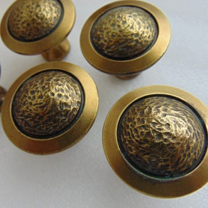 May include: A set of five round cabinet knobs with a metallic brass finish. Each knob features a smooth, flat outer rim and a raised, textured center with a hammered, irregular surface pattern that creates a distinct contrast in light and shadow.