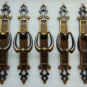 May include: Five antique bronze cabinet pulls. Each pull features a rectangular backplate with a textured center, a decorative top, and a curved handle. The pulls are approximately 6 inches long.