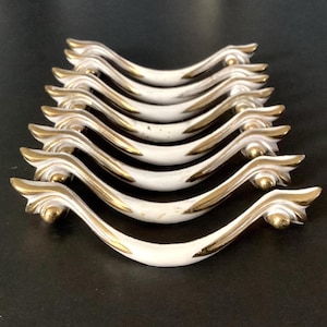 May include: A set of eight white and gold cabinet handles. The handles have a curved design with gold accents on the ends and along the edges. The handles are arranged in a row on a dark surface.