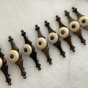May include: A set of eight drawer pulls featuring an elongated, dark bronze-toned metal backplate with a decorative, pointed shape. Each pull includes a circular, off-white ceramic knob centered on the metal plate with a small bronze-colored inlay.