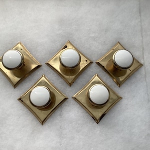 May include: A set of five cabinet knobs, each featuring a circular white ceramic center mounted on a square, metallic gold-toned brass backplate with pointed corners. The knobs are arranged in a cluster, showcasing the reflective finish of the metal.