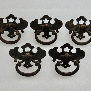 May include: Five antique bronze drawer pulls. Each pull features a decorative, ornate backplate with a curved handle. The pulls have a dark, aged finish, suggesting vintage or reclaimed hardware. The design is symmetrical and classic.