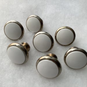 May include: A set of six circular buttons, each featuring a smooth, solid white center inlay surrounded by a polished metallic silver-toned rim. The buttons have a raised, rounded profile and a shank back for attachment to garments or fabric items.