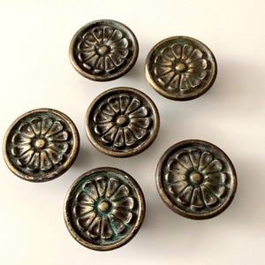 May include: Six antique bronze cabinet knobs with a floral design. The knobs have a round shape and a weathered, aged appearance. The bronze finish has hints of green patina, adding to their vintage charm.