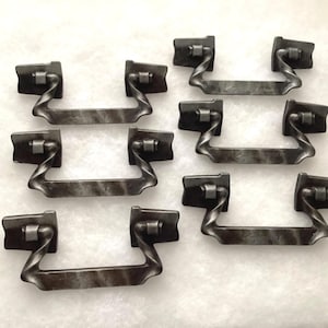 May include: A set of five matching dark gray metal drawer pulls. Each pull features a rectangular mounting plate on each side with a curved, twisted handle that connects to the plates. The metal has a mottled, textured finish throughout.