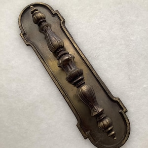 May include: A long, vertical brass drawer pull featuring a decorative, multi-segmented handle with a dark, aged patina. The handle is mounted on a matching rectangular brass backplate with rounded, notched corners and a recessed center border.