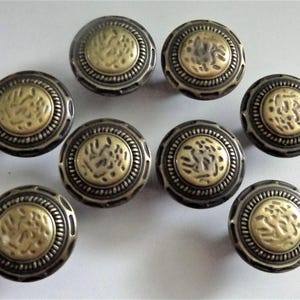May include: A collection of eight antique-style cabinet knobs. Each knob features a circular design with a brass-colored center and a dark, metallic outer ring. The center has a textured pattern, and the outer ring has a detailed, ridged edge.