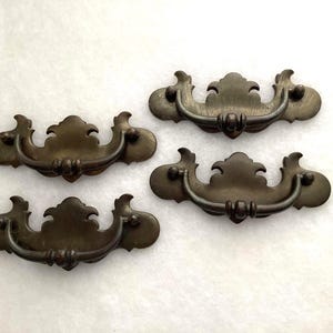 May include: A set of three matching drawer pulls in a dark bronze finish. Each pull features a decorative backplate with curved edges and a central crest, paired with a swinging bail handle attached by two small circular mounting points.