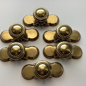 May include: A collection of six antique brass cabinet knobs. Each knob features a round, raised center and a decorative backplate. The knobs are arranged on a white surface, showcasing their aged, golden finish. Ideal for vintage furniture restoration.