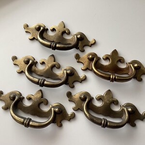 May include: Five antique brass drawer pulls with ornate, scroll-like designs. Each pull features a curved handle and decorative end pieces. The brass has a warm, aged patina, suggesting vintage or retro style. These pulls are suitable for furniture restoration.