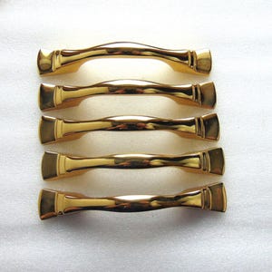 May include: Five gold-colored cabinet handles arranged horizontally. Each handle has a curved design with rectangular end pieces. The handles are made of a shiny, reflective material, suggesting a polished finish.