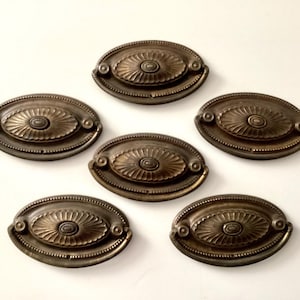 May include: Six antique bronze oval drawer pulls. Each pull features a central floral design surrounded by a detailed border. The pulls are arranged on a white surface, showcasing their aged, metallic finish.