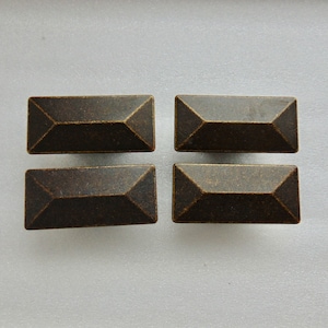 May include: Four rectangular cabinet pulls with a bronze finish. Each pull has a faceted design, with a slightly raised center and beveled edges. The hardware is arranged in a square formation against a neutral background.