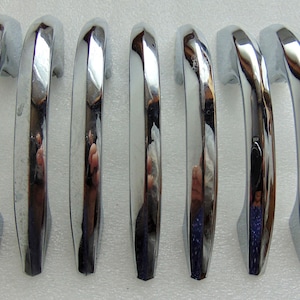 May include: Seven chrome-plated door handles arranged in a row. Each handle has a curved, elongated design with a shiny, reflective surface. The handles are likely for a vehicle or vintage furniture, and are set against a white background.