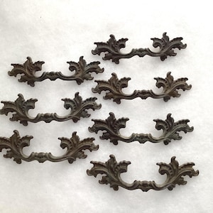 May include: A set of six dark brown metal drawer pulls arranged in two columns. Each pull features an ornate, symmetrical design with curved, leaf-like flourishes extending from the center mounting points to the outer edges of the handle.