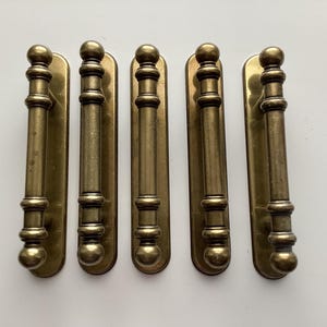 May include: Five antique brass door pulls. Each pull has a long, rectangular backplate with a cylindrical handle. The handles are detailed with decorative rings and ball finials. The brass has a warm, aged patina.