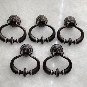 May include: A set of five dark bronze-toned metal drawer pulls. Each pull features a circular mounting plate and a swinging, teardrop-shaped handle with a central decorative accent ring. The hardware has a smooth, matte finish throughout.