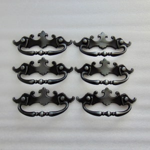 May include: Six antique-style drawer pulls with a dark, metallic finish. Each pull features a decorative backplate with a central flourish and a curved handle. The hardware is designed for furniture restoration or adding vintage charm.