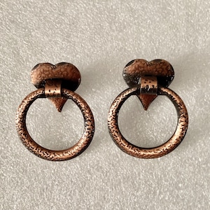 May include: Two antique copper-colored drawer pulls. Each pull features a heart-shaped top, a decorative ring, and a triangular accent. The pulls have a textured finish and are designed for furniture. The hardware is made of metal.
