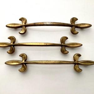 May include: Three antique brass drawer pulls with a fleur-de-lis design. The pulls are curved with a central bar and decorative ends. The brass has a warm, aged patina, suggesting vintage or antique hardware.
