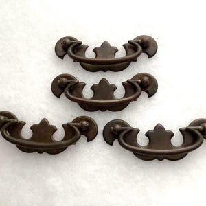 May include: Four matching dark bronze-toned metal drawer pulls. Each pull features a curved, swinging handle attached to a decorative backplate with a central pointed crest and rounded side accents, designed for furniture hardware applications.