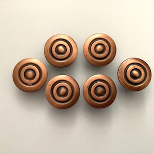 May include: Six antique copper-colored cabinet knobs arranged on a light background. Each knob is round with concentric circles and a central raised dot. The knobs have a vintage, aged appearance.
