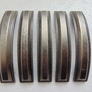 May include: Five antique-style cabinet pulls with a brushed, metallic finish. Each pull has a curved, rectangular shape with vertical grooves and a beaded edge. The hardware is a muted silver color, suitable for furniture restoration or new builds.