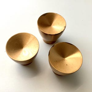 May include: Three gold-colored cabinet knobs with a conical design. The knobs have a rounded base and a flat, circular top. The finish has a slight patina, adding character to the hardware. These knobs are suitable for furniture or cabinetry.