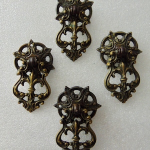 May include: Four antique bronze-colored drawer pulls with ornate, symmetrical designs. Each pull features a decorative top with a central handle and a stylized, openwork body. The pulls are arranged against a white background.