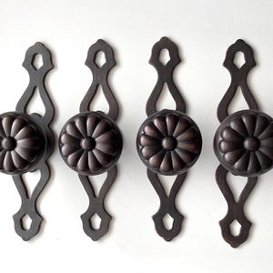 May include: Four dark bronze door pulls with a floral design on the round knob. The pulls have a decorative, elongated backplate with a diamond-shaped cutout at the top and bottom.