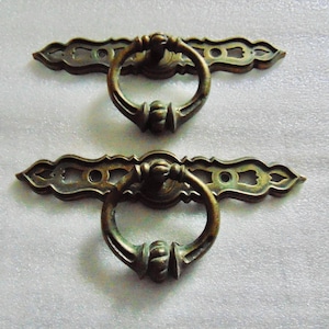 May include: Two antique bronze drawer pulls. Each pull features a decorative backplate with a ring handle. The bronze finish has a vintage look. The pulls are designed for furniture restoration or adding a classic touch to cabinets.