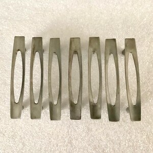 May include: Seven silver-toned cabinet pulls with an elongated oval cutout in the center. The pulls are rectangular with rounded edges and are arranged horizontally against a white background.