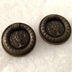 May include: Two antique bronze drawer pulls. Each pull features a circular design with a central emblem of a decorative urn and a surrounding ring handle. The pulls have a vintage aesthetic, suitable for furniture restoration or adding a classic touch.