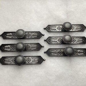 May include: A set of six dark gray metal drawer pulls, each featuring an elongated, pointed-end backplate with intricate cut-out patterns and a central, circular knob with a textured, rope-like border around the edge of the knob face.
