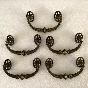 May include: Five antique bronze drawer pulls. Each pull has a curved handle with decorative detailing and starburst-shaped end pieces. The pulls are arranged on a white surface.