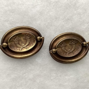 May include: Two oval-shaped brass drawer pulls featuring a central embossed design of two cornucopias filled with flowers. Each pull has a hinged handle attached to a decorative backplate with a smooth, metallic finish and a warm golden tone.
