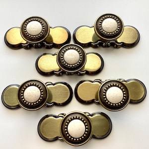 May include: Six antique brass cabinet pulls with white speckled ceramic centers. Each pull has a circular knob surrounded by a beaded edge, and a decorative brass backplate. These vintage hardware pieces are ideal for furniture restoration projects.