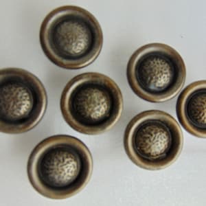 May include: Seven antique bronze cabinet knobs. Each knob features a textured center surrounded by a smooth, circular rim. The knobs are arranged on a white surface, showcasing their aged, metallic appearance. Ideal for furniture restoration or adding vintage charm.