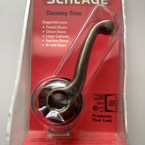 May include: A Schlage Dummy Trim door handle in a package. The handle is bronze-colored and has a curved design. The packaging is red and white, with text that reads "Dummy Trim" and "Products That Last."