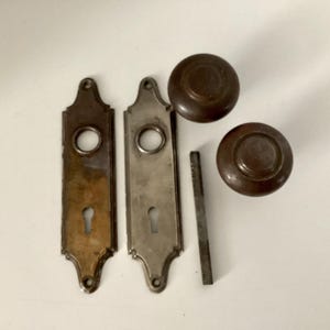 May include: Antique door hardware set, including two ornate backplates, two round doorknobs, and a threaded rod. The backplates are rectangular with keyholes and circular openings. The doorknobs and backplates have a bronze finish.
