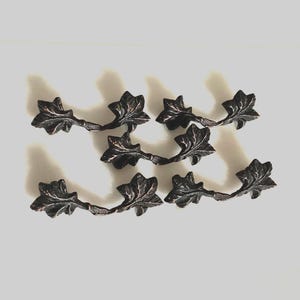 May include: Six bronze-colored leaf-shaped drawer pulls. Each pull features three detailed leaves connected by a curved stem. The hardware is designed for furniture and adds a decorative touch.
