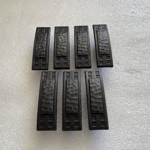May include: Seven black metal drawer pulls with a hammered finish and a square design. The pulls have a rectangular shape with a raised center section and a border of small squares.